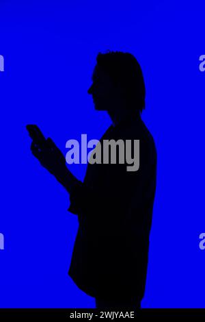 Silhouette un homme d'affaires élégant utilisant un smartphone moderne dans une veste debout sur un fond bleu brillant, un concept futuriste Banque D'Images