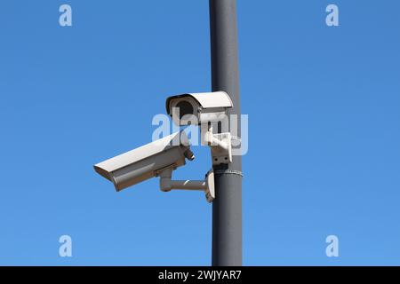 Deux petites fausses caméras de sécurité CCTV en circuit fermé blanc moderne factice regardant dans des directions opposées montées sur un poteau en métal gris foncé sur bleu clair Banque D'Images