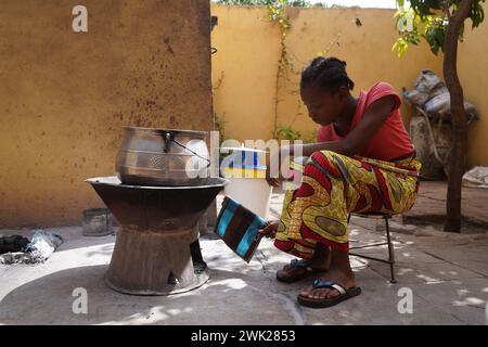 Jeune fille africaine assise devant un simple poêle à charbon de bois attisant le feu ; préparation traditionnelle des aliments Banque D'Images