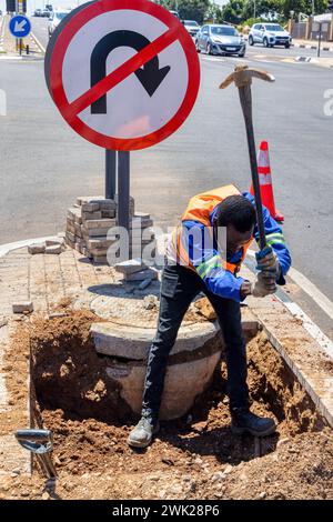 travailleur afro-américain avec une pioche creusant un fossé dans l'autoroute pour réparer le câblage sur une île à une intersection Banque D'Images