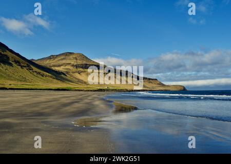 Plage de sable noir avec des rochers et des réflexes en rocade en Islande avec falaises, océan et herbe verte pendant une journée ensoleillée Banque D'Images