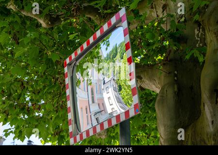 Un miroir de circulation rectangulaire avec un cadre rouge et blanc et un reflet déformé d'une ville. Le miroir est monté devant un arbre avec leav vert Banque D'Images