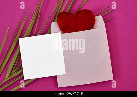 Maquette de carte d'invitation de mariage et enveloppe rouge avec jouet de coeur rouge et feuille de palmier sur fond rose. Maquette de carte vierge Banque D'Images