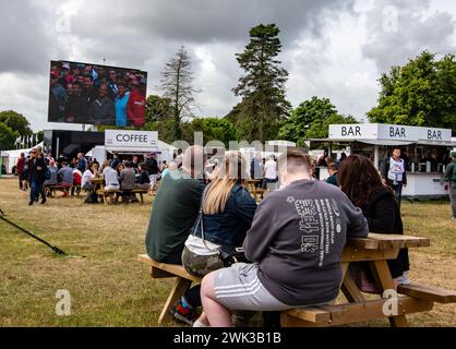 Les gens se sont assis autour de bancs avec des fourgonnettes de restauration servant des boissons alimentaires et regardant un grand écran au Festival de Goodwood de Speed West Sussex UK Banque D'Images