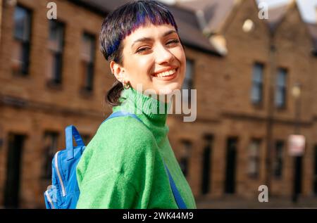 Jeune femme souriante en pull vert avec un sac à dos bleu marchant autour de la vieille ville sur une journée d'automne ensoleillée dame insouciante de se sentir en bonne santé et Banque D'Images