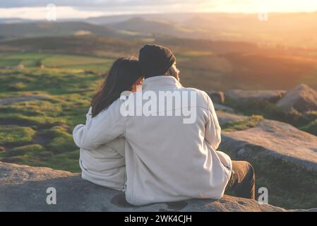 Vue arrière du couple heureux dans l'amour assis au sommet d'une montagne appréciant une vue de paysage de coucher de soleil image tonifiée Banque D'Images