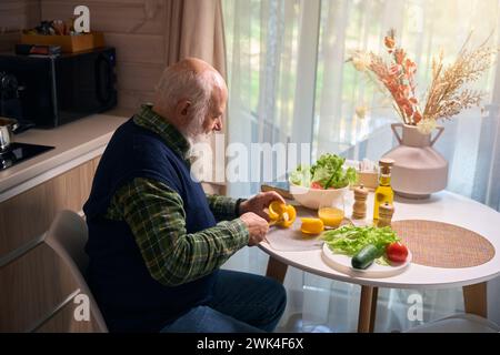 Un homme âgé à la table de la cuisine coupe des légumes pour la salade Banque D'Images