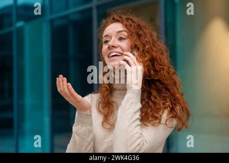 Portrait de belle jeune femme heureuse diplômée avec les cheveux roux bouclés parlant à un ami sur le mobile Banque D'Images