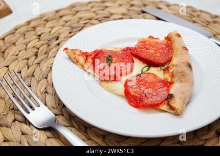 Une seule tranche de pizza pepperoni au basilic frais sur une assiette blanche sur un napperon en osier. Banque D'Images