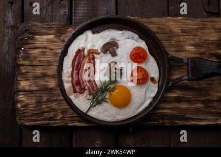 Oeufs frits avec bacon et légumes mis dans une poêle en fonte, sur fond de table en bois foncé vieux, Banque D'Images