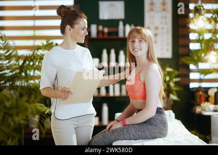 Temps de soins. thérapeute de massage médical féminin souriant dans l'armoire de massage avec presse-papiers et client adolescent faisant le check-up. Banque D'Images