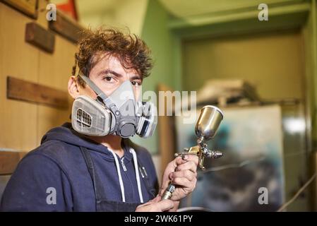 Portrait d'un jeune travailleur latin portant un masque de protection respiratoire regardant la caméra tenant un aérographe pendant le processus de peinture au pistolet Banque D'Images