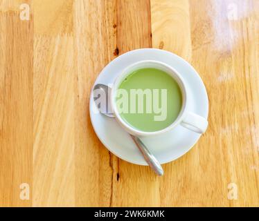 Vue de dessus du thé vert matcha au lait condensé chaud est servi dans une tasse blanche et une petite assiette avec une petite cuillère à thé sur fond de bois brun. Banque D'Images