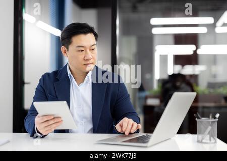 Homme asiatique concentré et sérieux travaillant dans le bureau à l'aide d'un cahier. Assis à un bureau et tenant une tablette. Banque D'Images