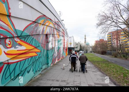 Les gens avec un chien marchent devant un graffiti mural coloré devant des bâtiments résidentiels, derrière eux une maison de machine à vapeur en forme de mosquée Banque D'Images