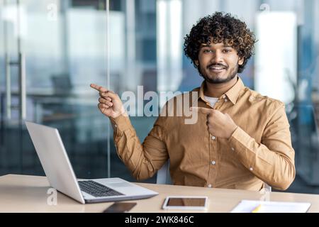 Jeune homme souriant indien assis dans le bureau à un bureau avec un ordinateur portable, regardant la caméra et pointant ses doigts sur le côté. Banque D'Images