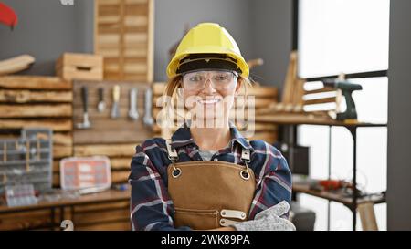 Attrayante jeune femme blonde charpentier, lunettes de sport et casque rigide, frappe une pose confiante avec les bras croisés dans son atelier de menuiserie intérieure. Banque D'Images