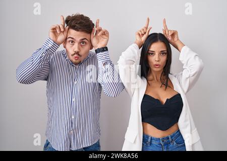 Jeune couple hispanique debout sur fond blanc faisant un geste drôle avec le doigt sur la tête comme cornes de taureau Banque D'Images