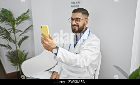 Un homme barbu souriant dans un manteau blanc prend un selfie dans une pièce moderne avec une plante et un signe wi-fi Banque D'Images