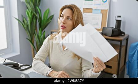 Une femme hispanique réfléchie examine des documents dans un bureau moderne, évoquant professionnalisme et concentration. Banque D'Images