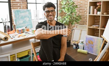 Un jeune homme hispanique avec une barbe souriant dans un studio d'art rempli de peinture et de toiles. Banque D'Images