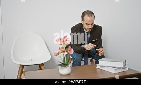 Homme hispanique chauve dans un costume assis à l'intérieur regardant des lunettes près d'une chaise blanche et des fleurs roses Banque D'Images