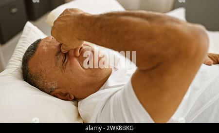 Homme hispanique se reposant dans la chambre, montrant de la détresse ou des maux de tête tout en étant allongé sur un lit blanc dans un cadre intérieur. Banque D'Images