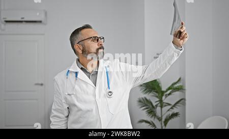 Bel homme hispanique mature avec une barbe grise, portant un manteau de médecin, examine une radiographie dans une salle de clinique lumineuse. Banque D'Images