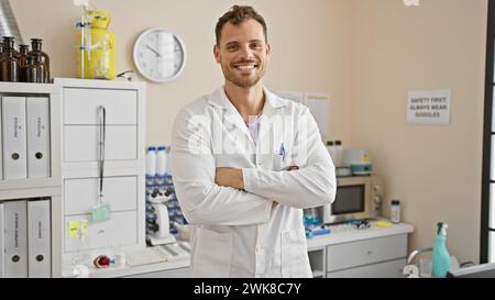 Beau jeune homme barbu dans une blouse de laboratoire se tient confiant avec les bras croisés dans une clinique d'hôpital. Banque D'Images