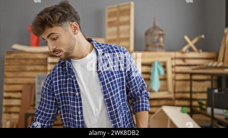 Beau, sérieux jeune homme arabe, un maître charpentier, debout dans son atelier de menuiserie bourdonnante, complètement entouré par le bois, t Banque D'Images