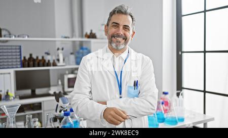 Souriant, confiant, jeune scientifique hispanique aux cheveux gris, debout avec un geste de bras croisés dans le labo Banque D'Images