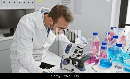Le scientifique hispanique Man analyse des échantillons à l'aide d'un microscope dans un cadre de laboratoire entouré de tubes à essai et de béchers. Banque D'Images