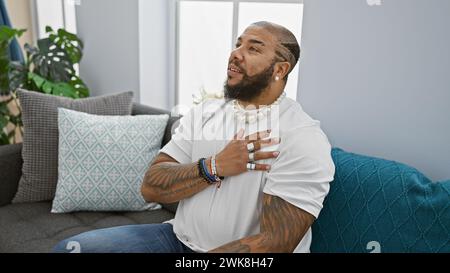 Bel homme barbu avec des tatouages plaçant la main sur le cœur dans un intérieur confortable du salon Banque D'Images