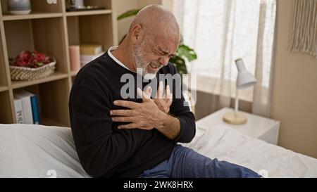 Homme chauve mature serrant la poitrine dans la douleur dans la chambre à coucher, exprimant la détresse et le besoin urgent d'aide médicale. Banque D'Images