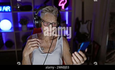 Technolo, une femme senior aux cheveux gris, passionne le monde du jeu vidéo en streaming, avec manette à la main, depuis sa salle de jeu éclairée par l'obscurité Banque D'Images