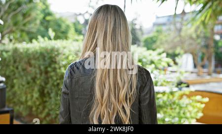 Une jeune femme se tient au milieu d'une verdure luxuriante dans un parc serein, ses cheveux blonds coulant sur une veste décontractée, illustrant la tranquillité urbaine. Banque D'Images