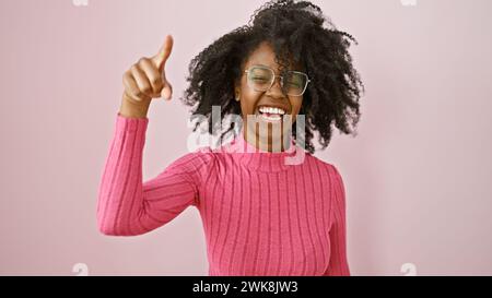 Une joyeuse femme afro-américaine portant des lunettes donnant un pouce vers le haut dans une pièce rose. Banque D'Images