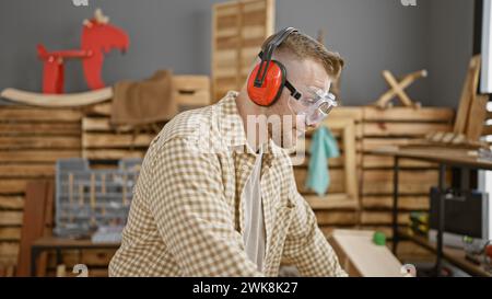 Bel homme à la barbe portant un équipement de sécurité travaillant avec du bois dans un atelier de menuiserie. Banque D'Images