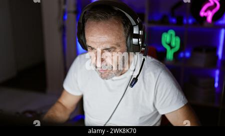 Homme aux cheveux gris d'âge moyen plongé dans une session de jeu intense sur son stream, immergé dans la lueur néon de sa salle de jeu Banque D'Images