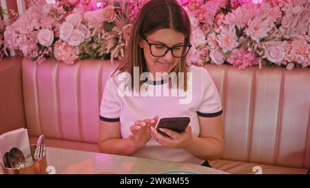 Femme souriante à l'aide d'un smartphone dans un café orné de fleurs roses Banque D'Images