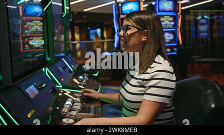 Une jeune femme brune souriante aime jouer aux machines à sous dans un cadre de casino dynamique. Banque D'Images