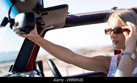 Une jeune femme caucasienne ajuste ses lunettes de soleil dans une voiture lors d'un voyage en voiture Banque D'Images