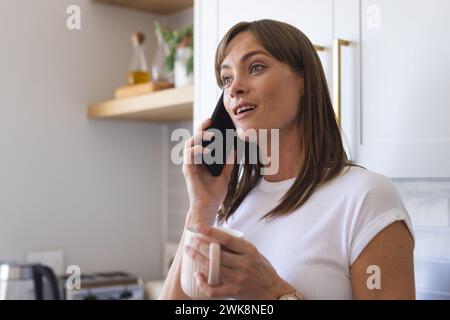 Une femme caucasienne d'âge moyen est engagée dans une conversation téléphonique tout en tenant une tasse Banque D'Images