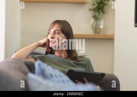 Une femme caucasienne d'âge moyen se détend sur un canapé, tenant une tablette et paraissant réfléchie Banque D'Images
