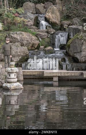 Londres, Royaume-Uni - 15 février 2024 - Un garçon se tient sur un pont de pierre devant une cascade et des falaises rocheuses dans le jardin de Kyoto, espace pour le texte, Focus sélectif. Banque D'Images