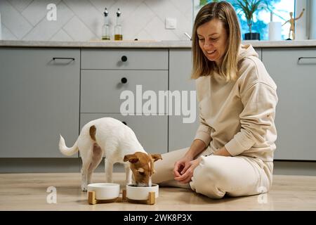 Femme mettant le bol de nourriture avec la nourriture pour son chien sur le sol dans la cuisine, propriétaire femme passant du temps avec l'animal de compagnie à la maison, alimentation animale et animal CA Banque D'Images