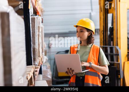 travailleuse dans le casque de sécurité et gilet de sécurité à l'aide d'un ordinateur portable tout en vérifiant l'inventaire dans l'entrepôt Banque D'Images