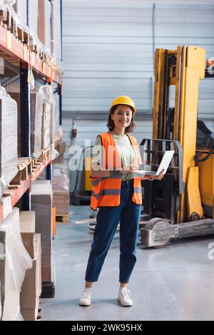 travailleur féminin d'entrepôt heureux dans le casque de sécurité et gilet de sécurité à l'aide de l'ordinateur portable tout en vérifiant l'inventaire Banque D'Images