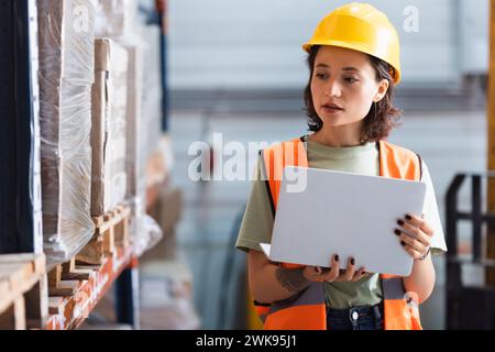 travailleuse dans le casque de sécurité et gilet de sécurité à l'aide d'un ordinateur portable tout en vérifiant l'inventaire dans l'entrepôt Banque D'Images