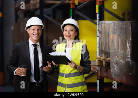 homme d'affaires souriant en costume et casque de sécurité tenant le café près de l'employé féminin, concept logistique Banque D'Images